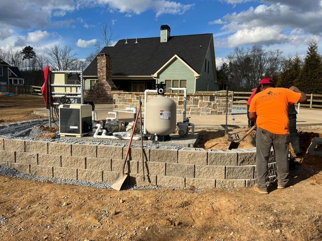 A man in an orange shirt is working on a brick wall in front of a house.