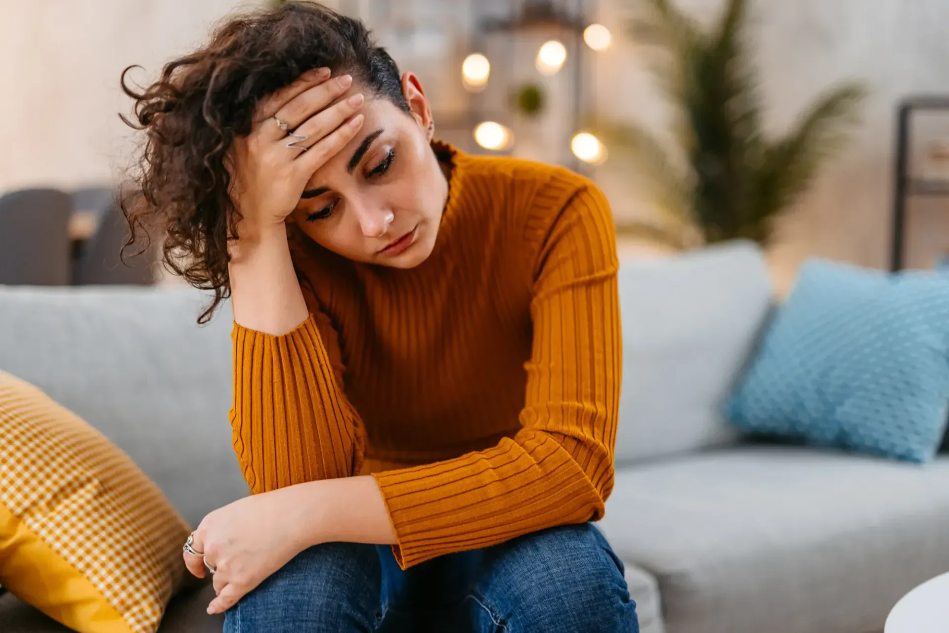 Woman with hand on forehead, looking down, seated on a couch indoors, appears stressed.