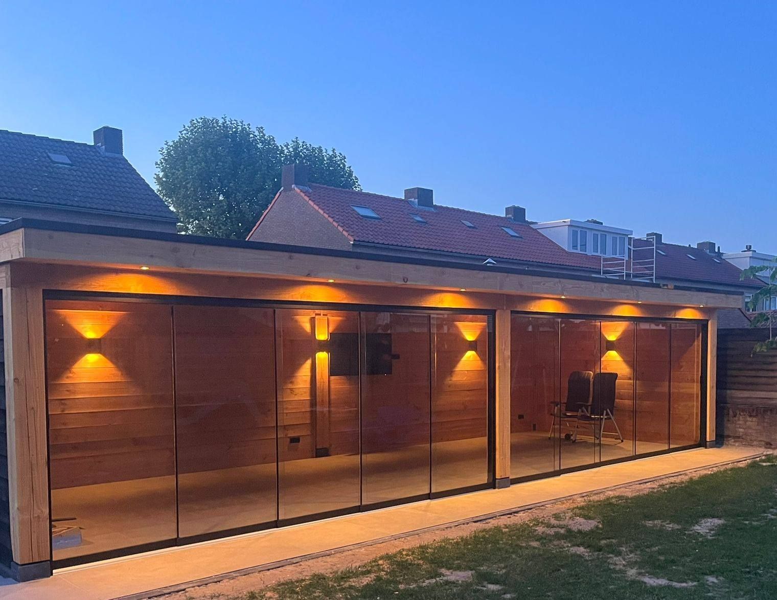 Terras met houten frame en glazen wanden, verlicht door warme lampen. Achtertuin bij zonsondergang.