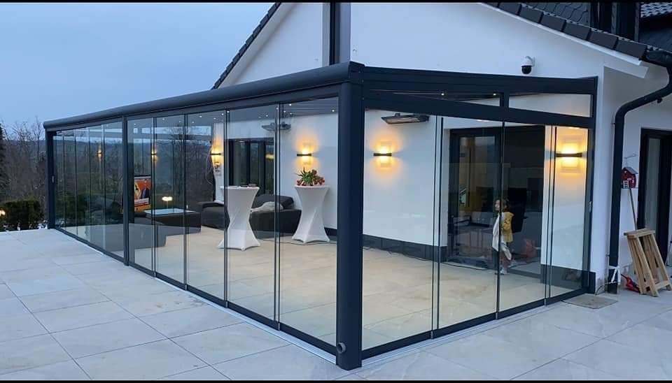 Een patio met glazen wanden, grenzend aan een wit huis, met tafels en warme verlichting.