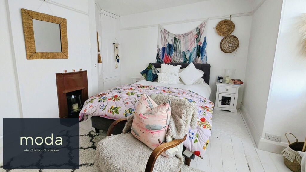 Bedroom with floral bedding, tapestry headboard, and a sheepskin chair. White walls, wood floors.