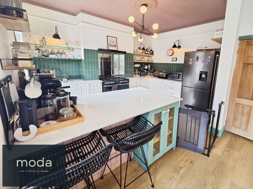 Kitchen with island and stools; green tile backsplash, black appliances, light wood floor, pink ceiling.