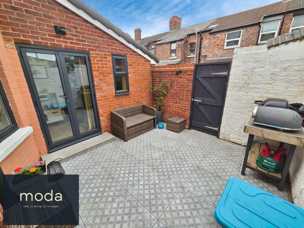 Patio area with gray tiled floor, brick walls, and black French doors. A wicker bench, small table, and grill are visible.