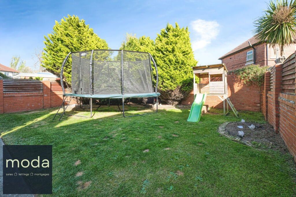 Backyard with trampoline, slide, and small flowerbed. Green grass, brick walls, and a blue sky.
