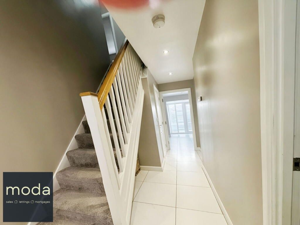 Hallway with staircase, neutral walls, white trim, tiled floor, and a glimpse of a light-filled space at the end.