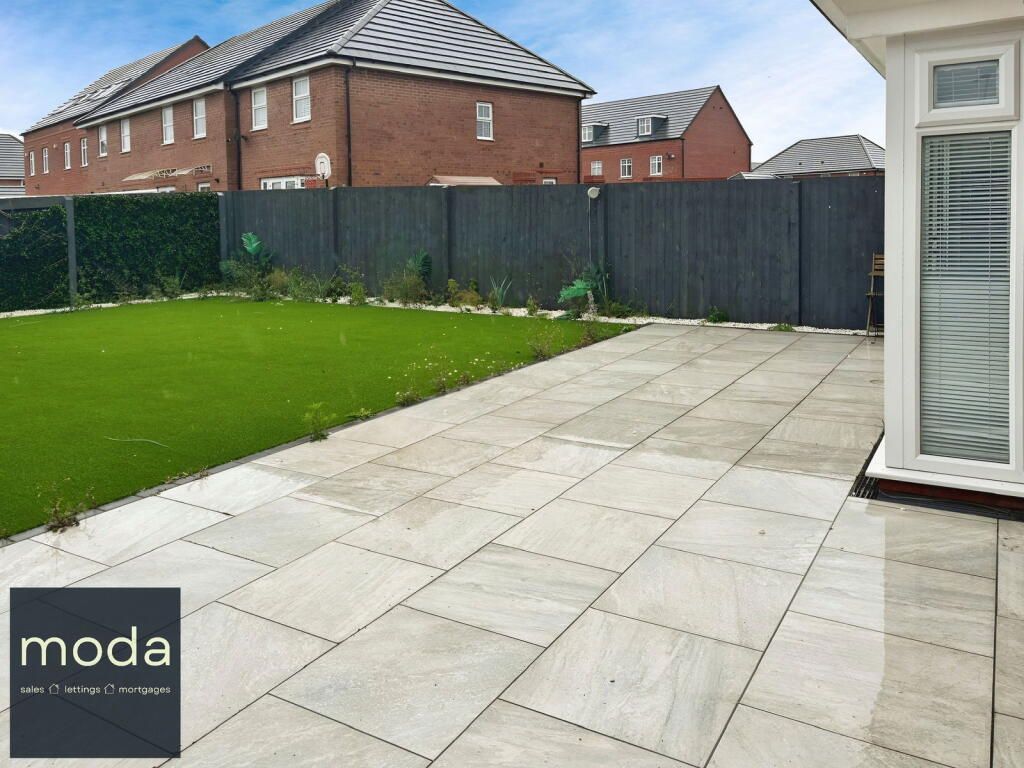 Patio with gray paving, green lawn, dark fence, and brick houses in the background.
