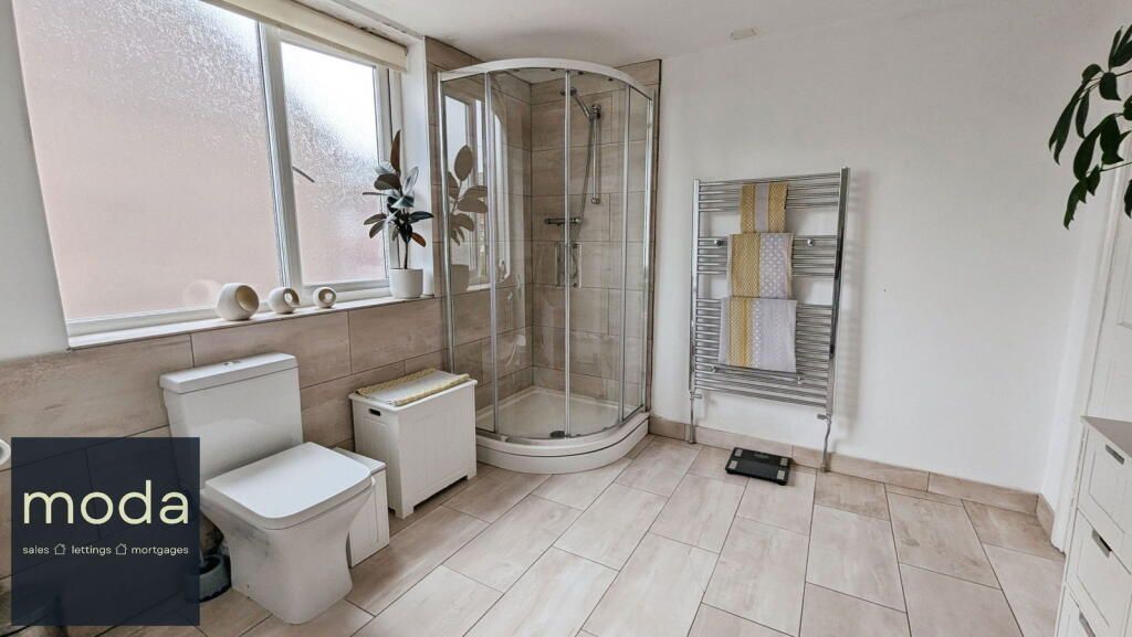 Bathroom with a shower, toilet, towel rack, and window; light-colored walls and flooring.