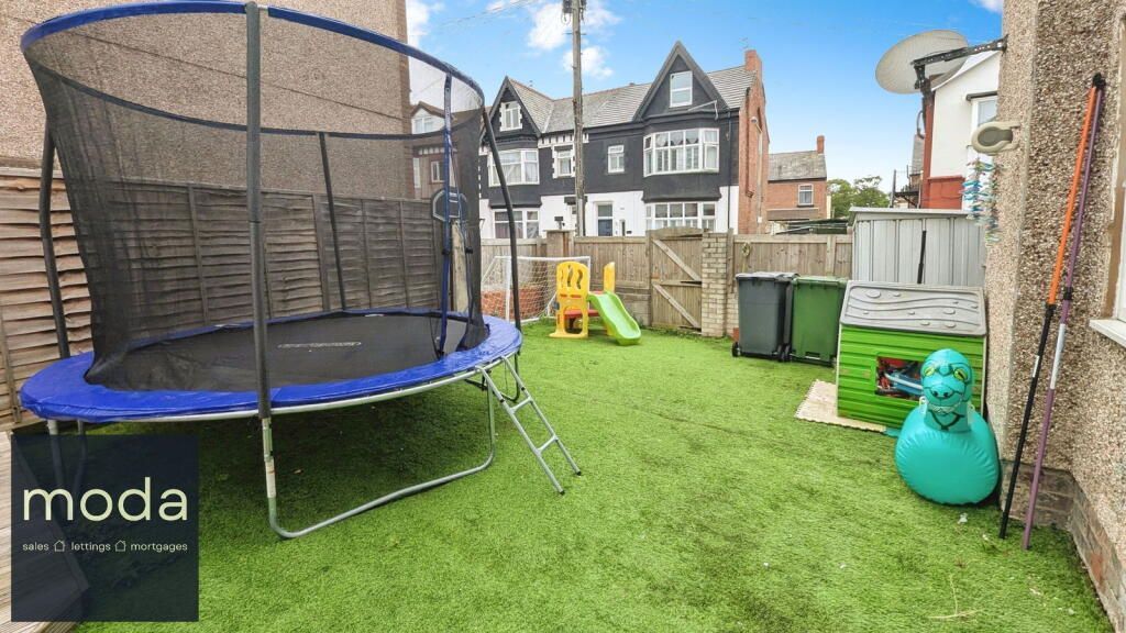 Backyard with trampoline, play slide, and lawn. Houses and blue sky in background.