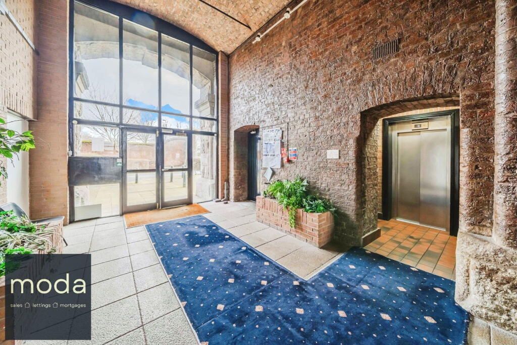 Lobby with glass doors, blue carpet, textured stone walls, and an elevator. Plants add some greenery.
