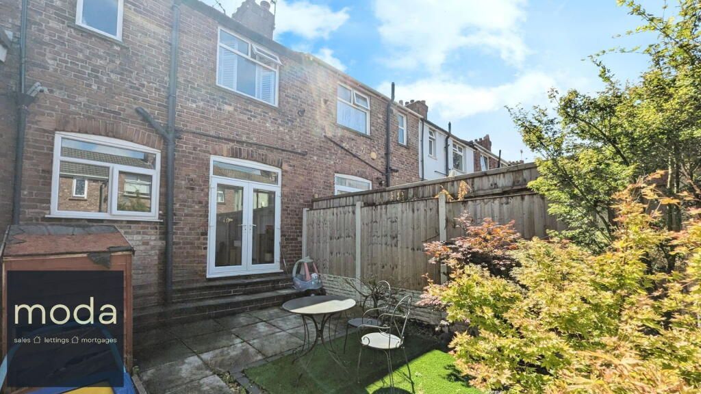 Backyard of a brick house with patio, small table, and shrubs under a blue sky.