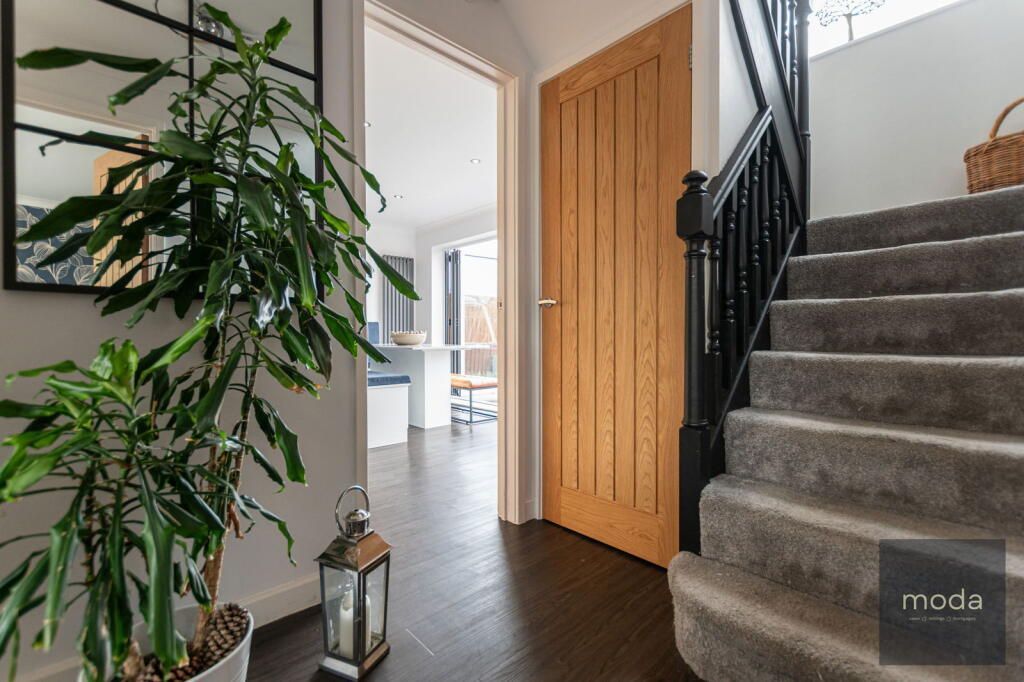 Hallway with staircase, wooden door, large plant, and mirror. Dark wood flooring and neutral walls.
