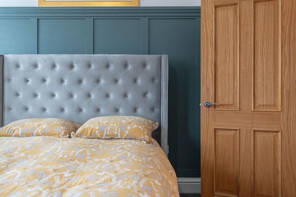 Bedroom with yellow bedding, a tufted gray headboard, and a teal wall with wood paneling, next to a wooden door.