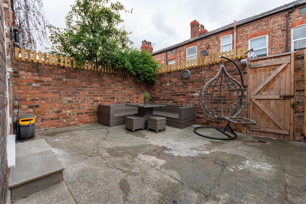 Brick-walled patio with a hanging chair, outdoor seating set, and wooden fence. Cloudy sky.