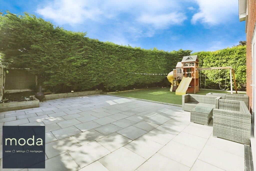 Backyard with patio, grass, playset, and tall green hedge against a blue sky.