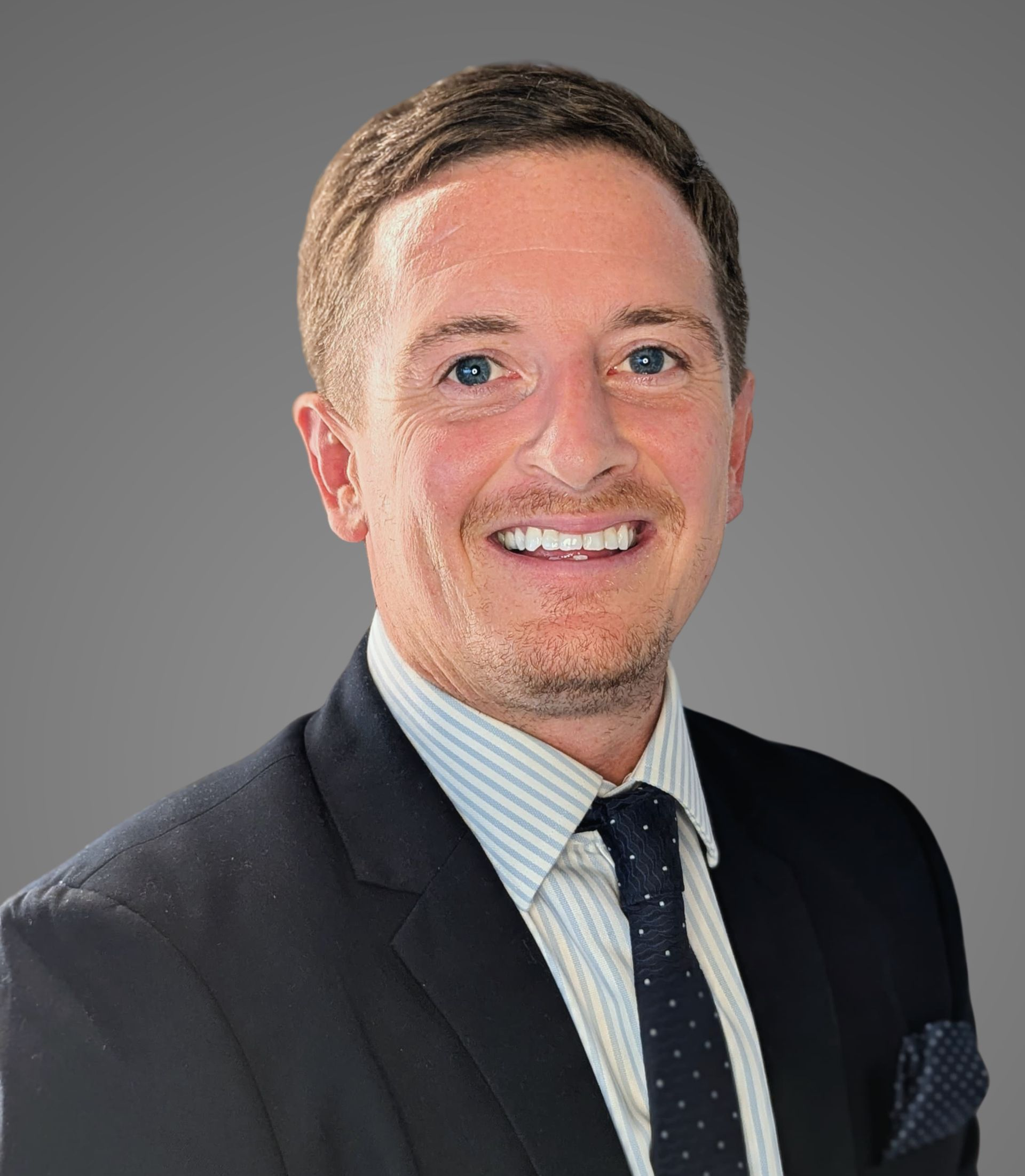 Man in suit smiling at the camera. Light skin, short brown hair, blue tie, grey background.