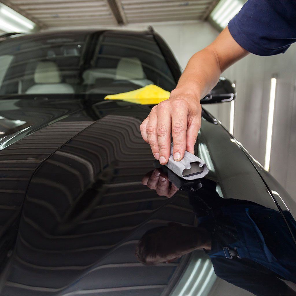 Person applying detailing clay to a black car hood in a well-lit shop.