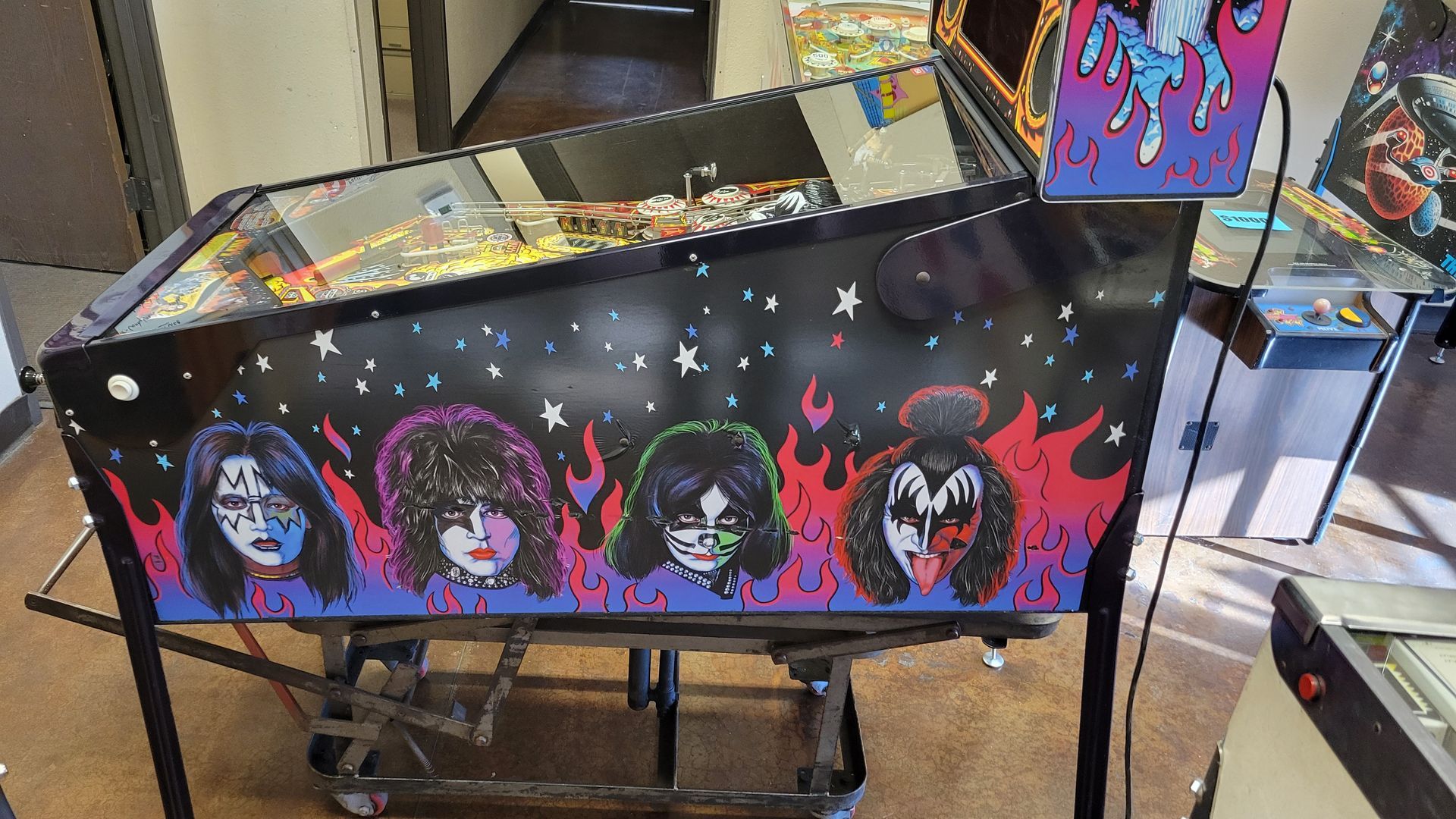 A kiss pinball machine is sitting on a cart in a room.
