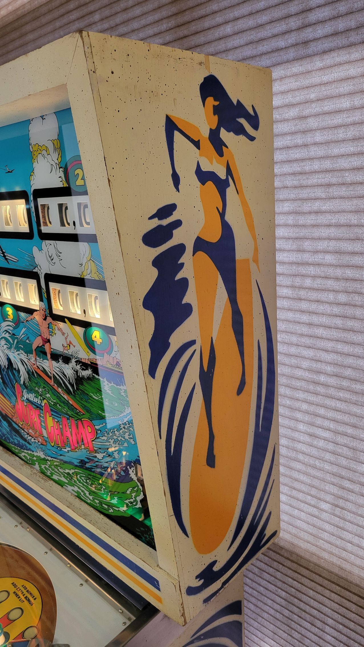 A woman in a bikini is standing on a surfboard on the side of a pinball machine.