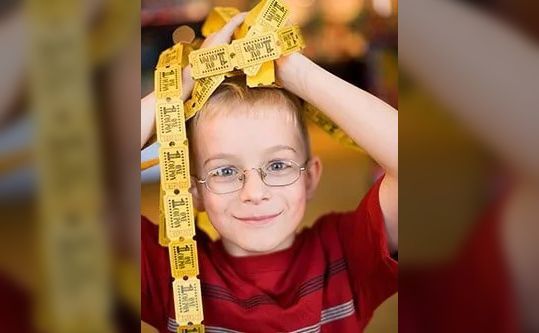 A young boy wearing glasses is holding a string of yellow tickets over his head.