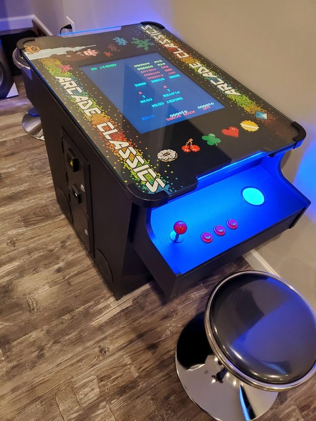 An arcade classics arcade machine is sitting on a wooden floor next to a stool.