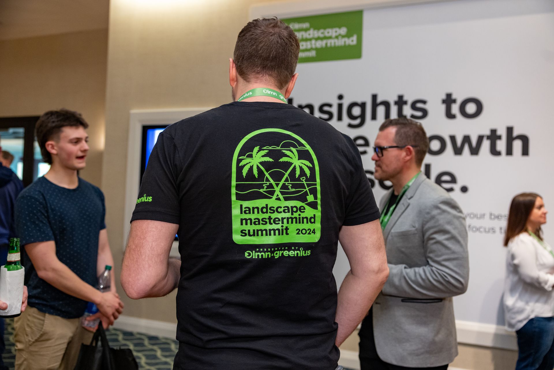 A man wearing a landscape management summit shirt is standing in a room with other people.
