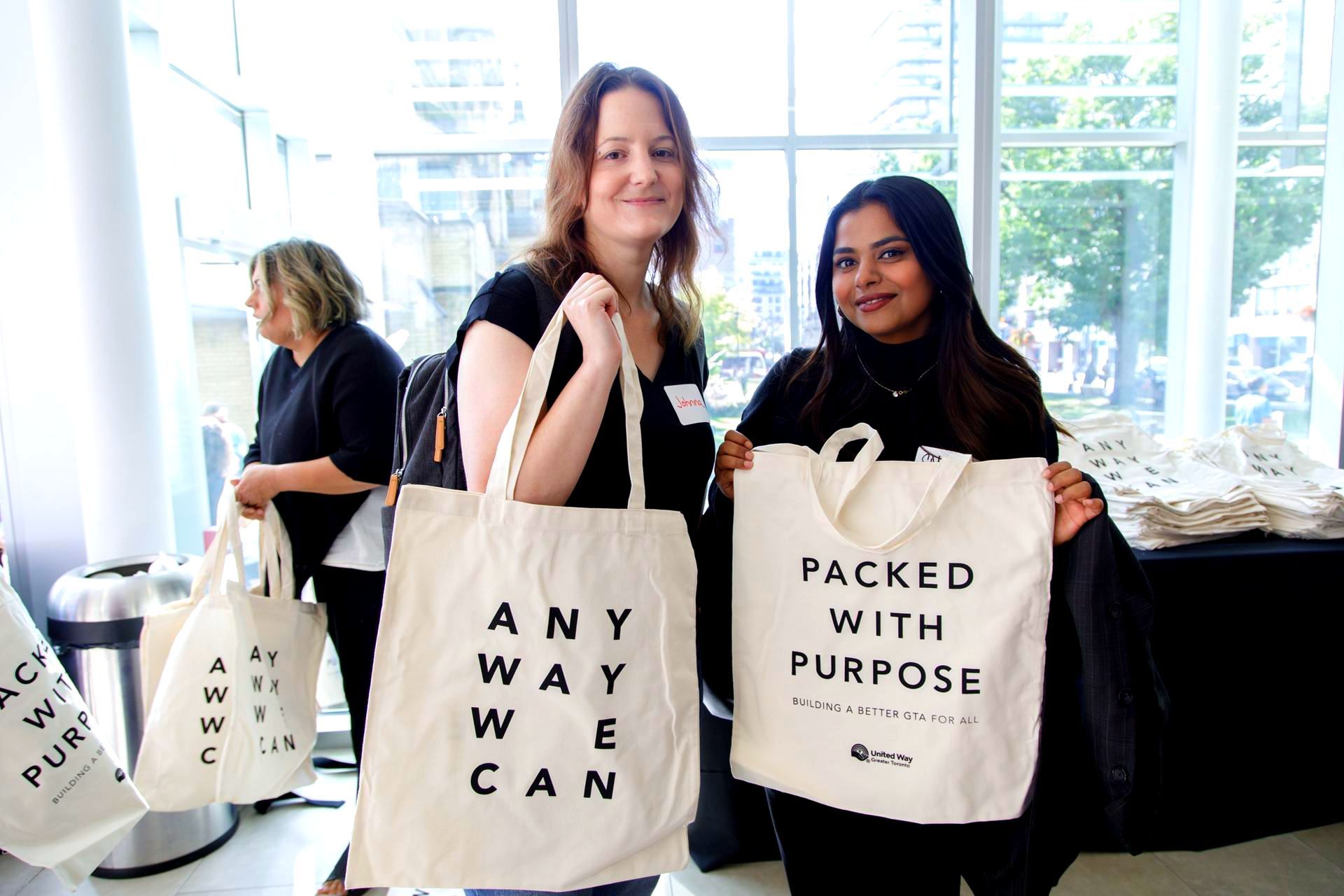 Two women are holding tote bags that say packed with purpose