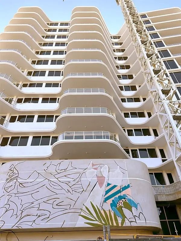 White high-rise building with curved balconies, mosaic mural at base, construction crane visible.