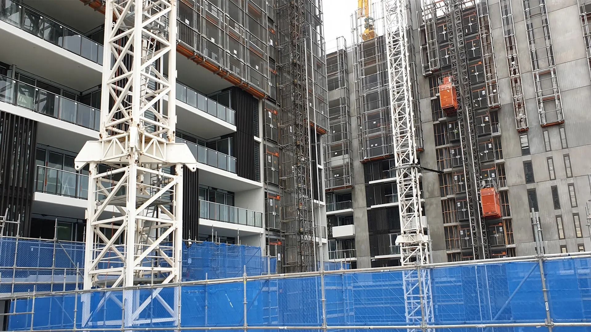 Construction site with multiple high-rise buildings; white cranes; blue safety nets.