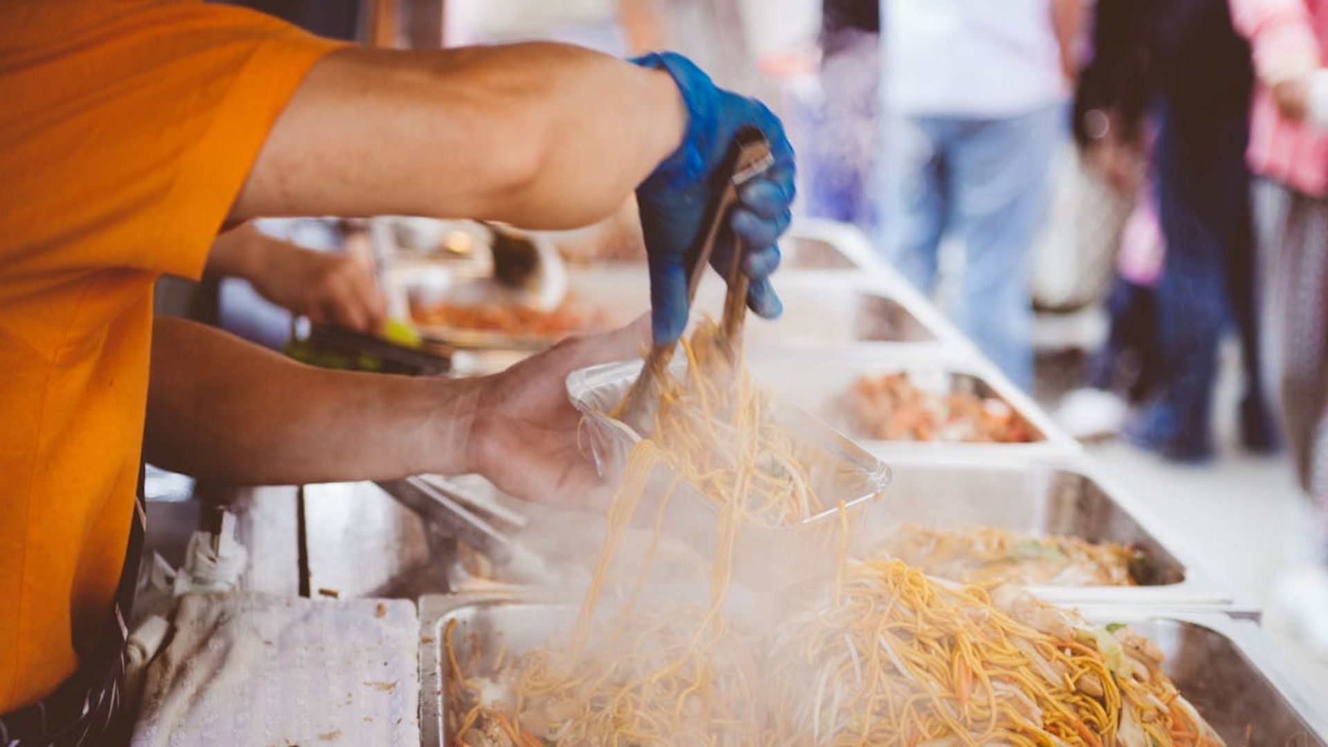 Una persona sirve fideos de un bufé. Lleva guantes azules en las manos y la comida humea.