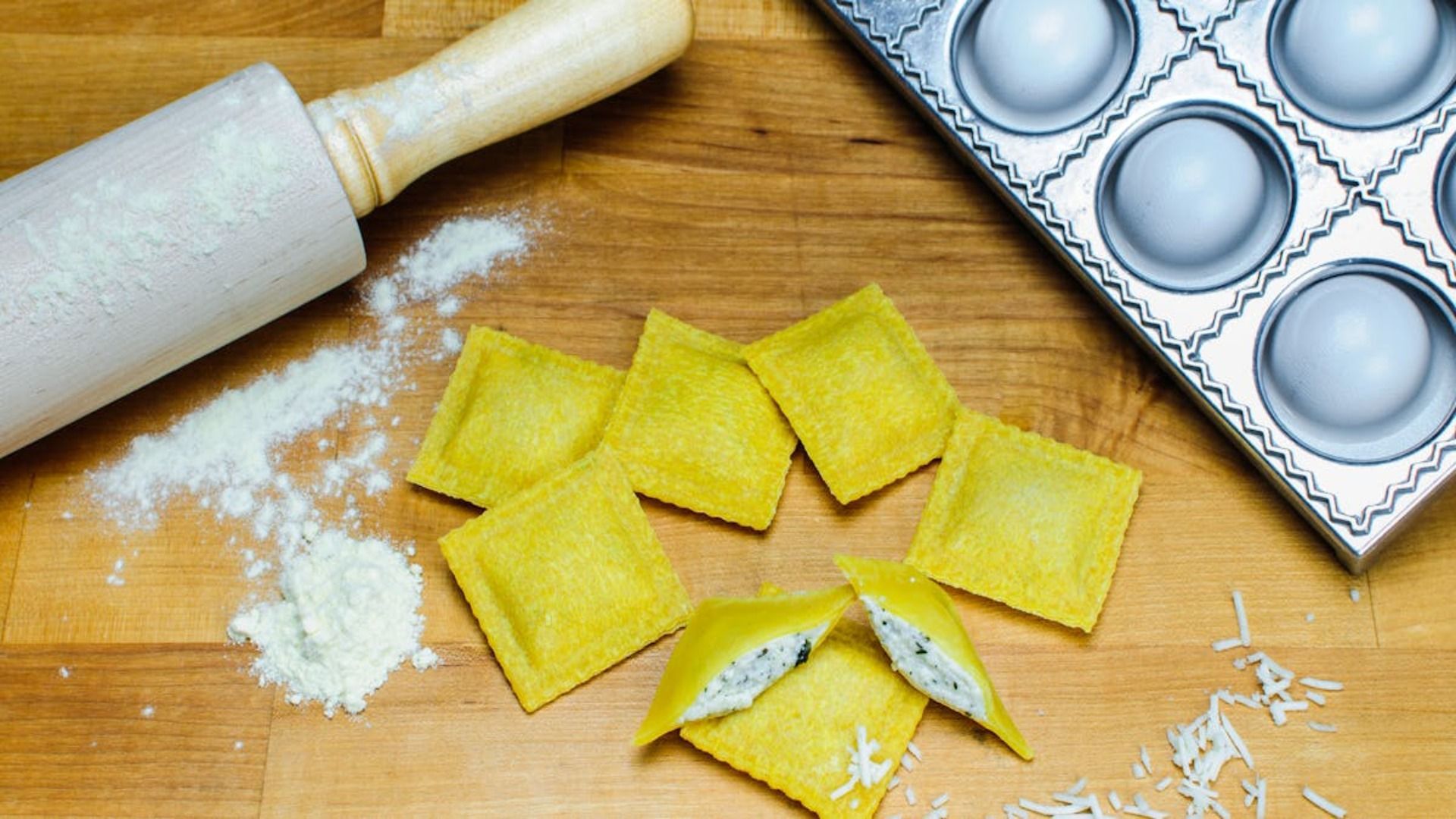 Escena de preparación de pasta: ravioles, rodillo, harina y un molde para ravioles sobre una superficie de madera.