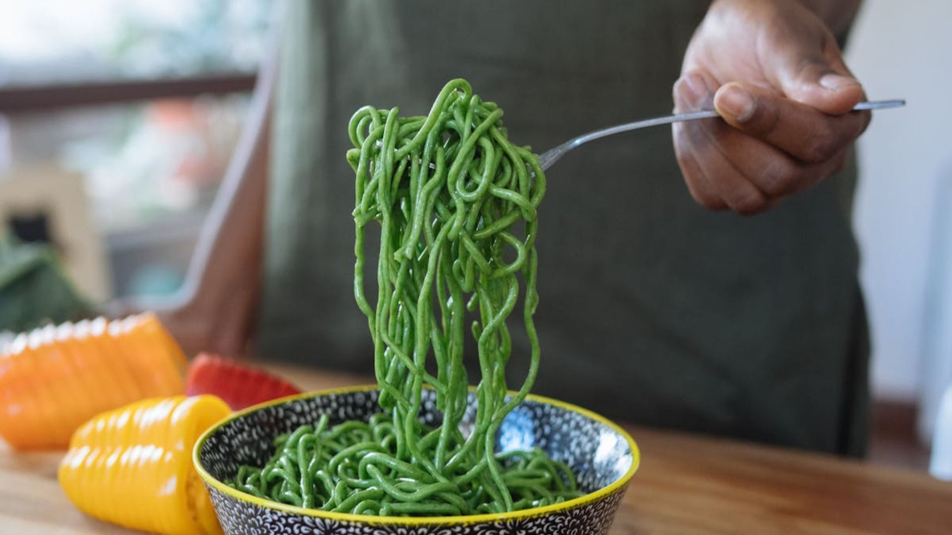 Persona que sostiene un tenedor con pasta de color verde brillante de un recipiente estampado, con pimientos morrones de colores cerca.