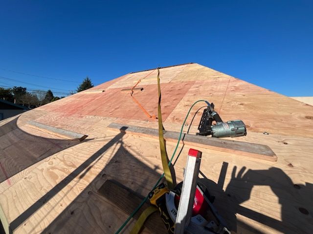 A work site showing a partially constructed roof with plywood, a pneumatic nailer, and safety gear under a clear blue sky.