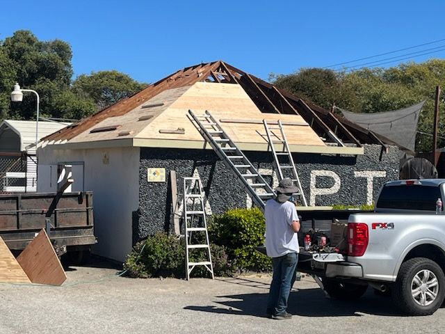 Construction worker standing near a partially demolished building with a ladder. Roof is exposed, truck parked nearby.