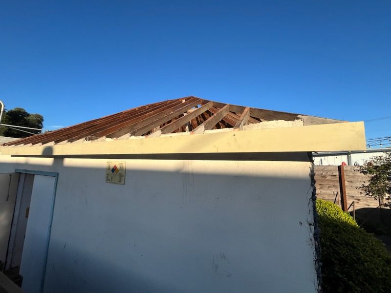 Building with a partially dismantled roof, revealing wooden beams. Cream-colored exterior walls, blue sky background.