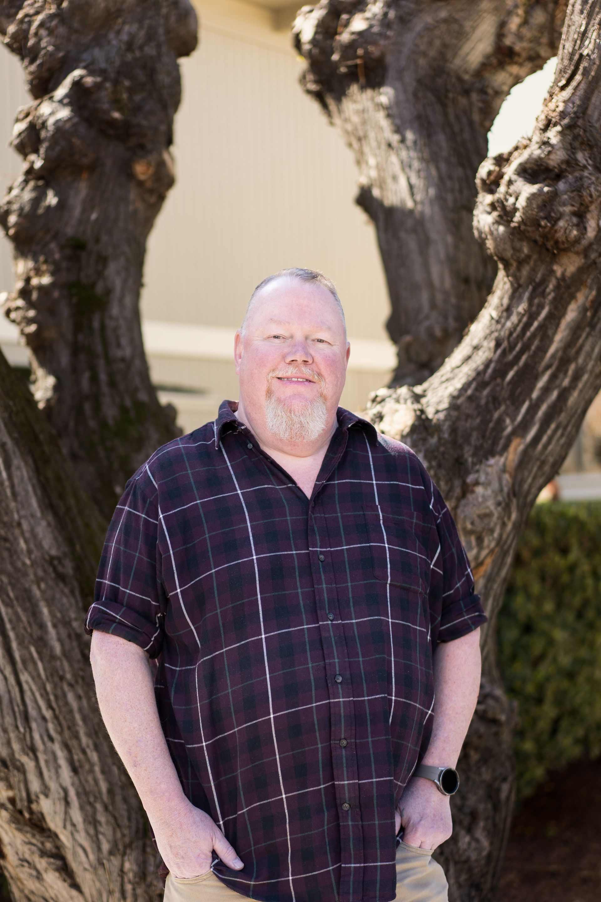 Man with red hair and beard, in plaid shirt, smiles outdoors. Leans on a tree.