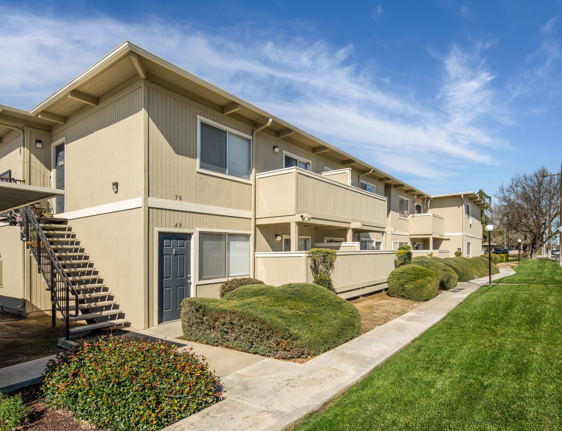 Two-story beige apartment building with stairs and balconies, set against a blue sky and green grass.