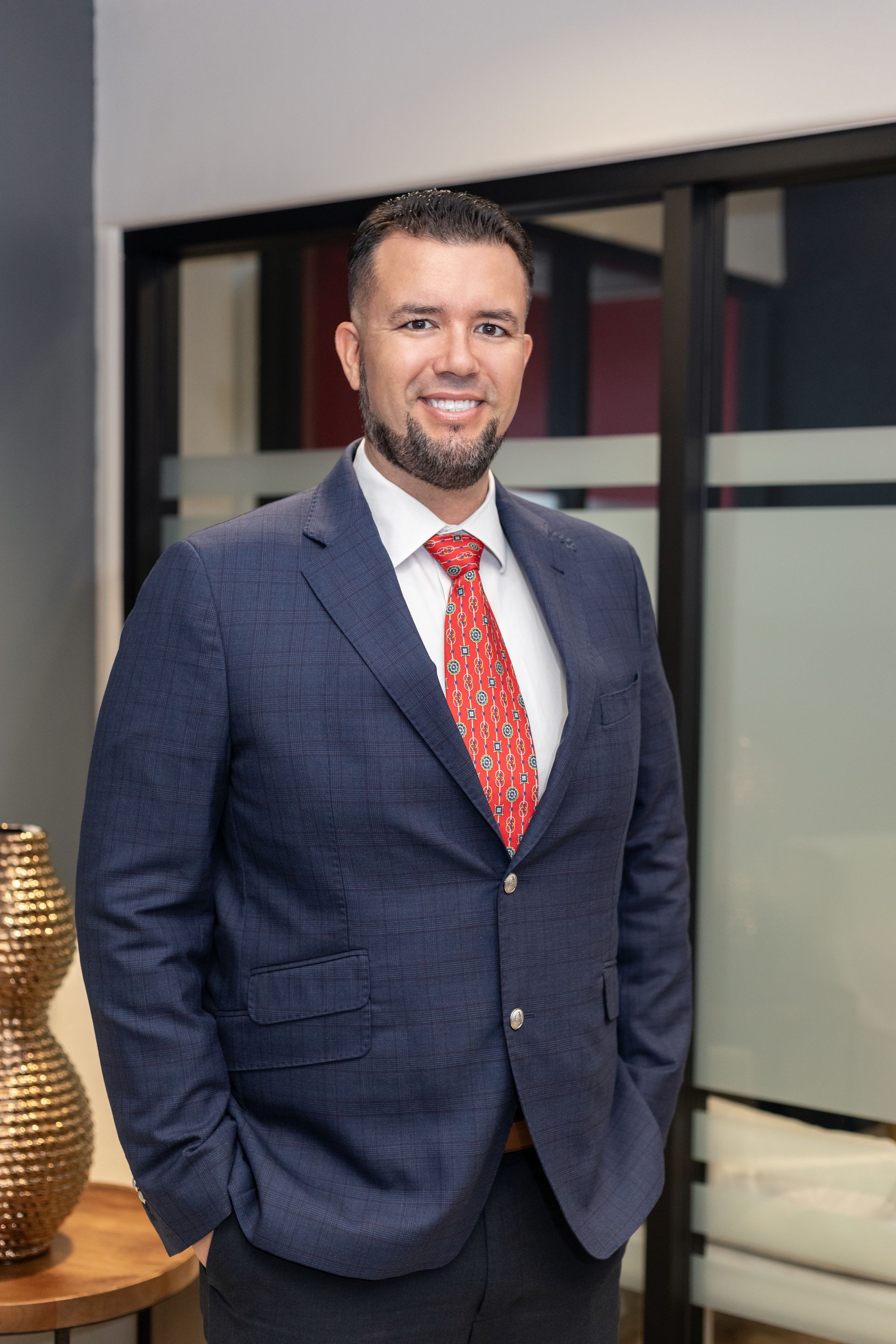 Man in blue blazer, red patterned tie, smiling, hands in pockets, near a glass door.