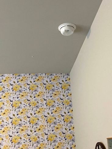 Corner of a room with floral wallpaper, white wall, gray ceiling, and a smoke detector on the ceiling.