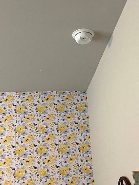 Corner of a room with floral wallpaper, white wall, gray ceiling, and a smoke detector on the ceiling.