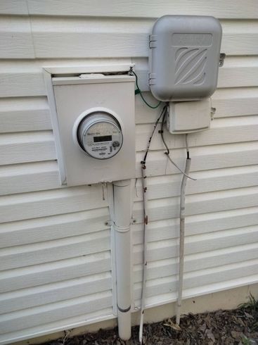 Electrical meter and utility boxes mounted on a white house exterior with wires and conduit.