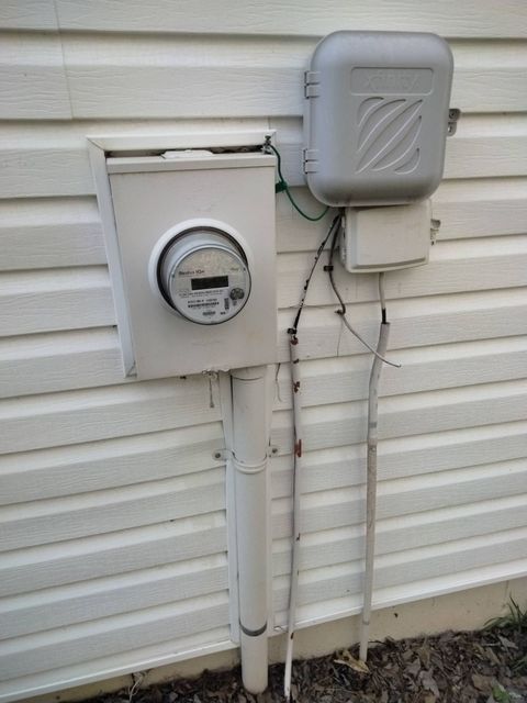 Electrical meter and utility boxes mounted on a white house exterior with wires and conduit.