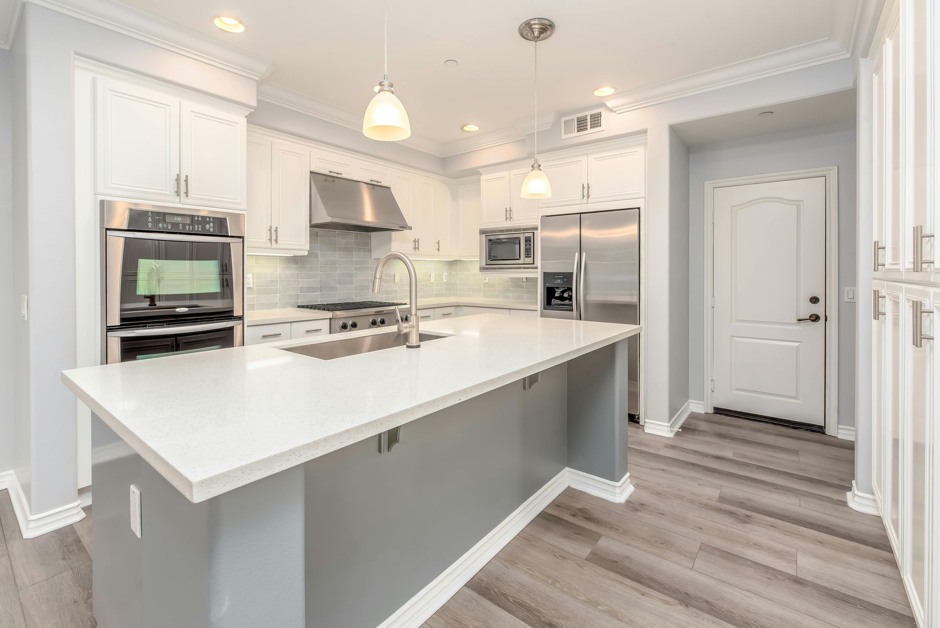 Bright modern kitchen with white cabinets, large island, stainless steel appliances, and gray wood flooring Bright modern kitchen with white cabinets, large island, stainless steel appliances, and gray wood flooring