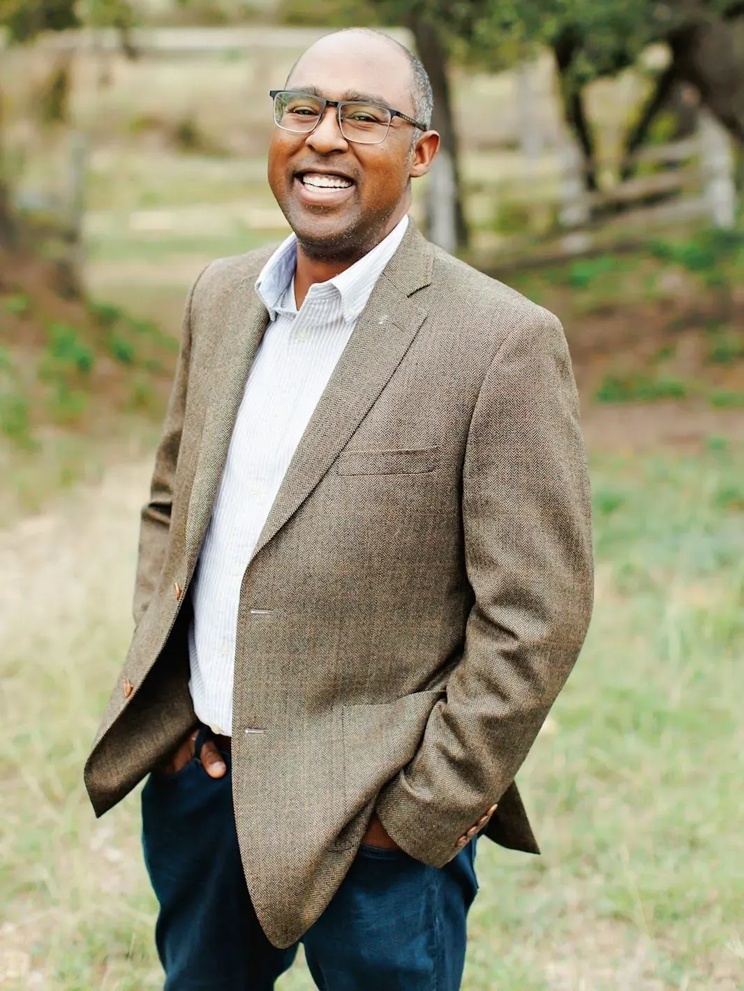 Man in a brown blazer, smiling with hands in pockets, standing outdoors.