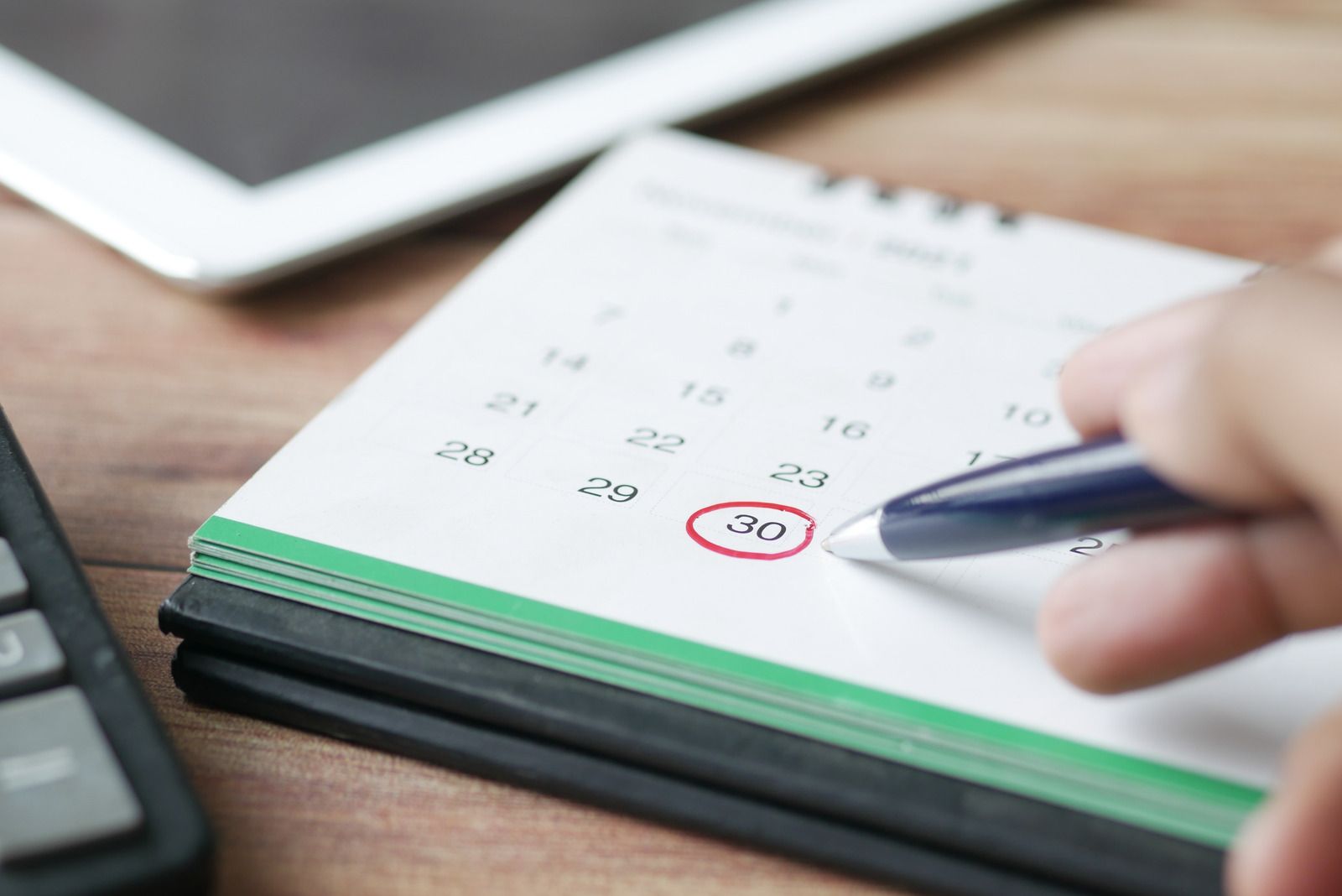 Person's hand circling date on calendar with a pen, next to a tablet and calculator.