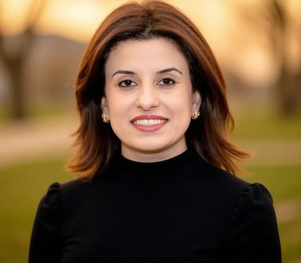 Woman with shoulder-length brown hair, smiling, wearing a black turtleneck and gold earrings, outdoors.
