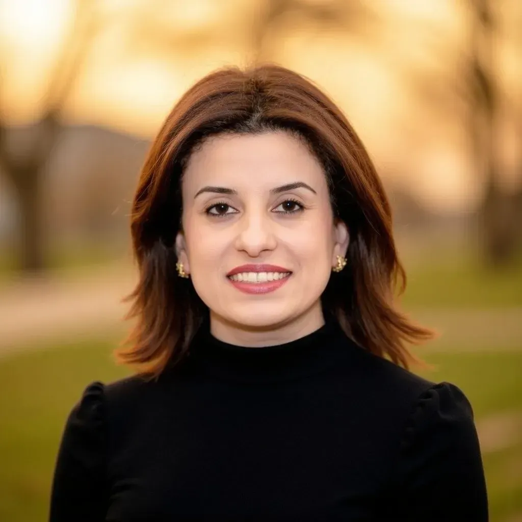 Woman with shoulder-length brown hair, smiling, wearing a black turtleneck and gold earrings, outdoors.