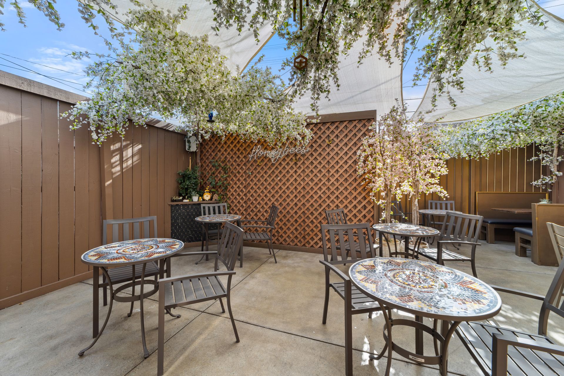 Outdoor patio with tables and chairs, under a canopy and decorative trees. Brown walls and a trellis in the background.