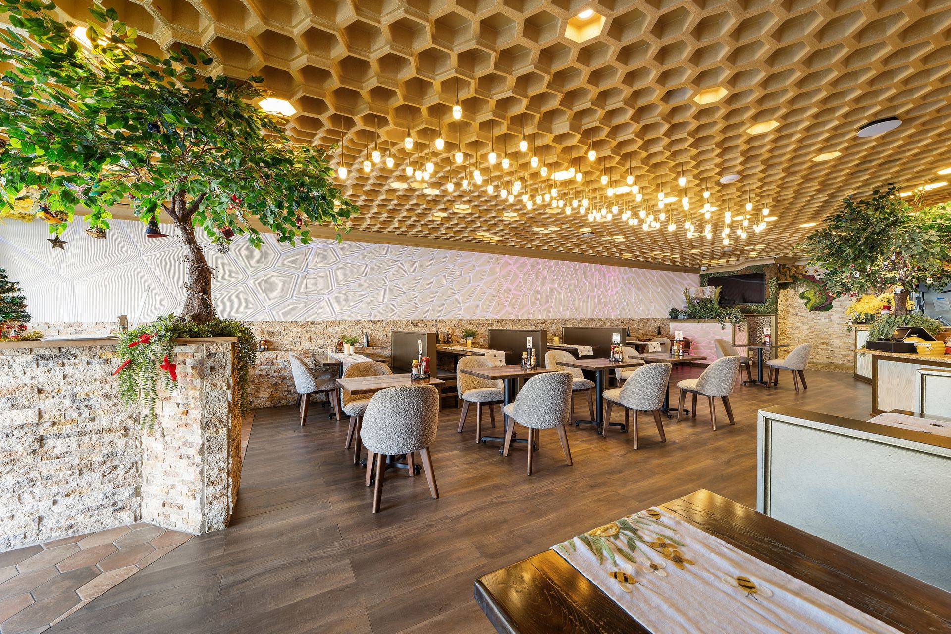 Restaurant interior with wooden floor, tables, chairs, artificial trees, and textured ceiling.