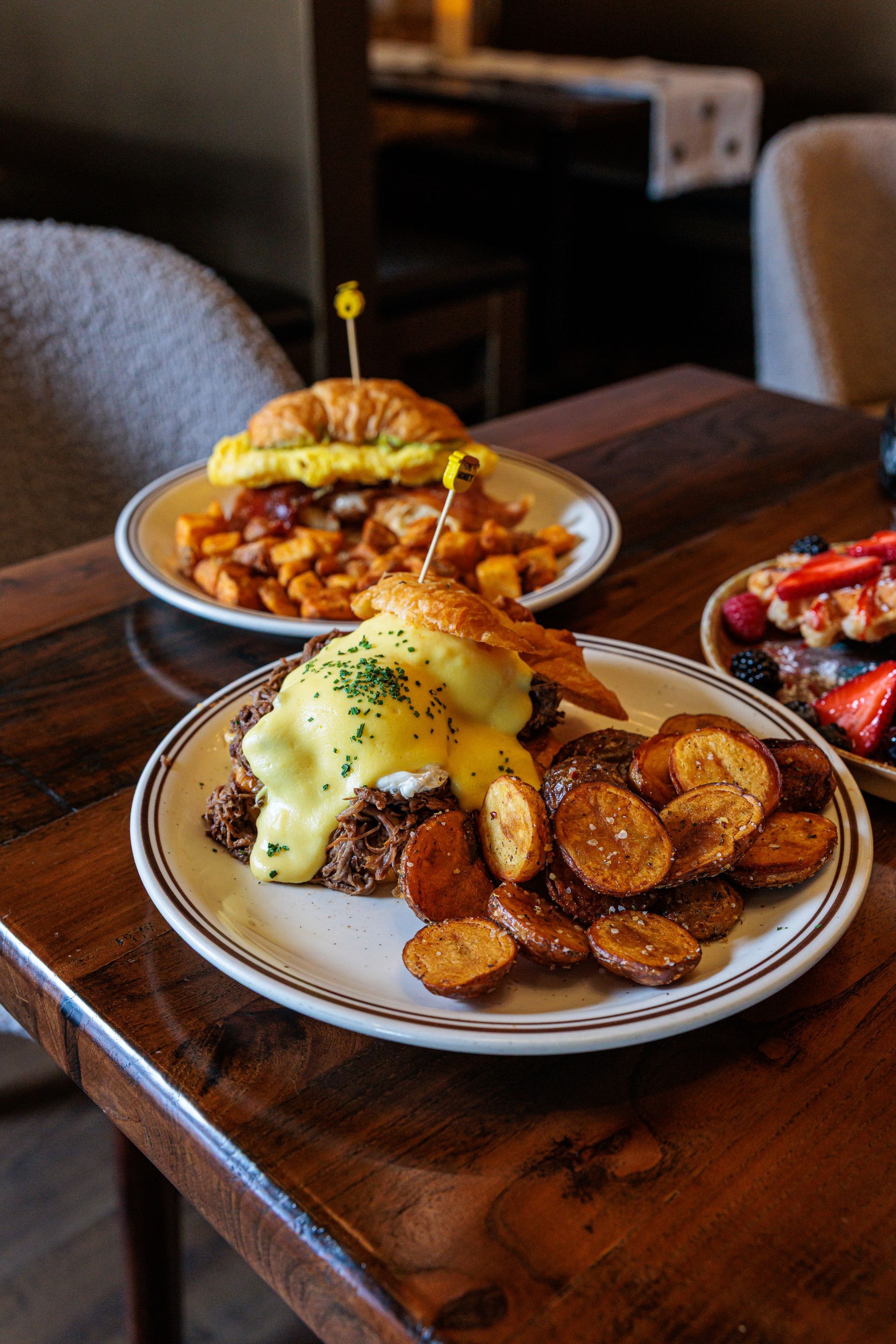 Eggs Benedict with potatoes and a breakfast sandwich on a wooden table.