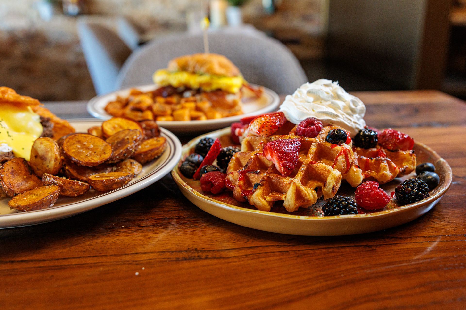 Breakfast dishes with waffles, berries, potatoes, and a breakfast sandwich on a wooden table.